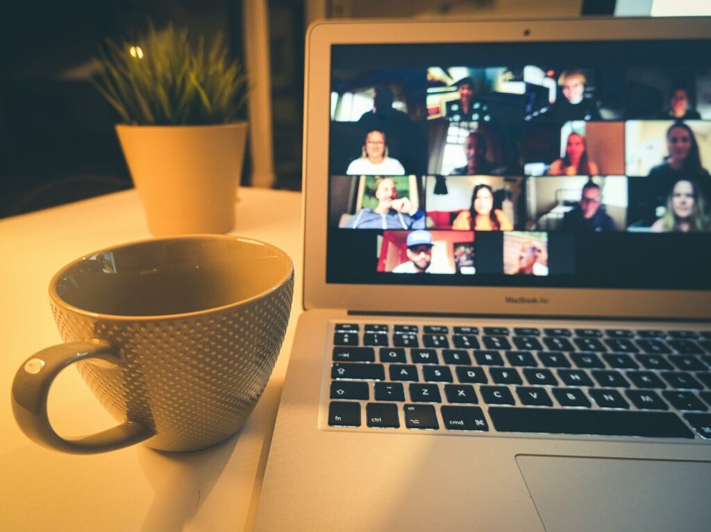 A laptop displaying a video conference with many people beside a coffee mug